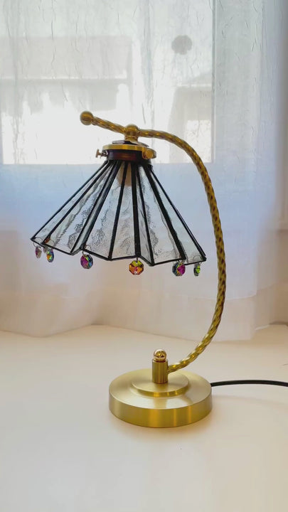 Vintage brass stained glass table lamp glowing warmly on a bedroom nightstand with cozy bedding and soft amber light.